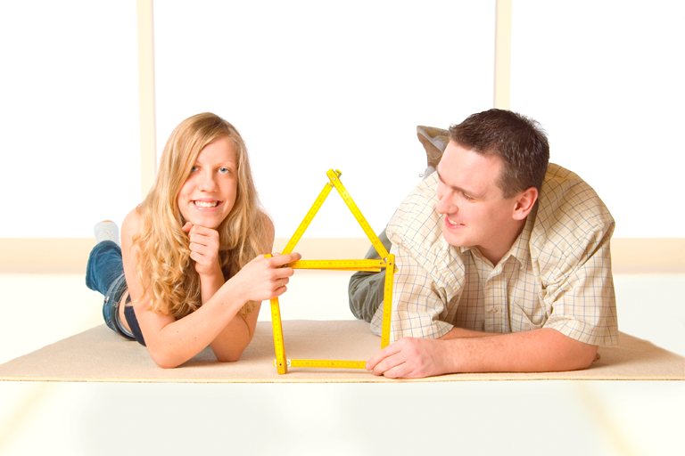 Pärchen auf Fußboden liegend in unmöblierter Wohnung hält ein Haus geformt aus einem Zollstock